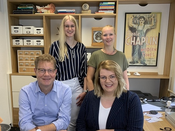 Vier Mitarbeitende eines modernen Büros sitzen und stehen vor Regalen und einem Charlie Chaplin Poster.